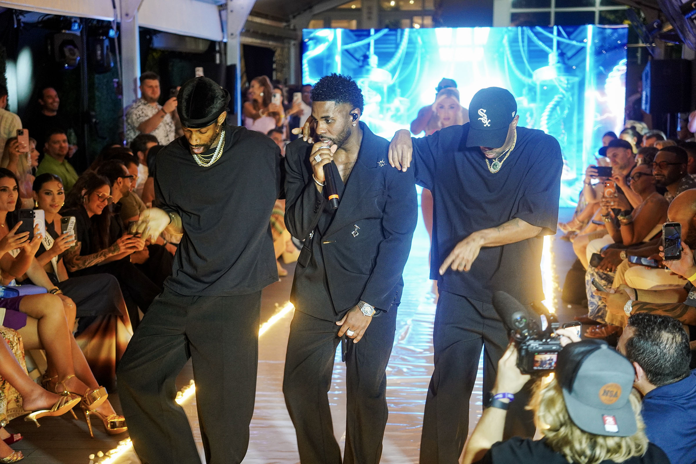 Jason Derulo performs with dancers as the runway audience and photographers surround the stage.